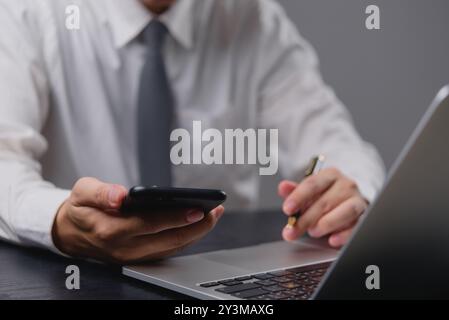 Businessman holding smartphone, using laptop. Managing tasks, modern office setup. Focus on digital communication, business operations. Professional w Stock Photo