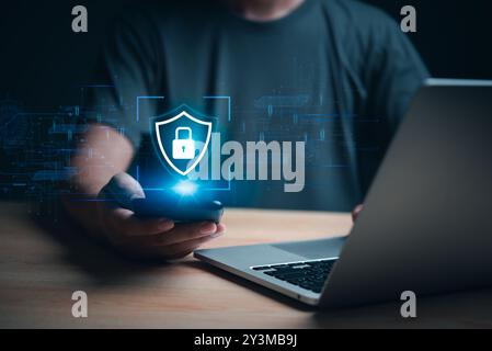 Hand holding smartphone with cybersecurity hologram, laptop in background. Represents data protection and digital security. Great for tech firms, cybe Stock Photo