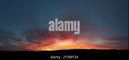 Stunning sunset paints sky with vibrant hues of red, orange, and pink, blending into deep blues and purples. Silhouetted hills create contrast against fiery horizon, adding drama to serene landscape. Stock Photo