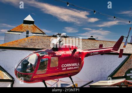 A red helicopter marked as Cornwall Air Ambulance is parked in front of a white building with a distinctive roof. The sky is bright blue with some clo Stock Photo