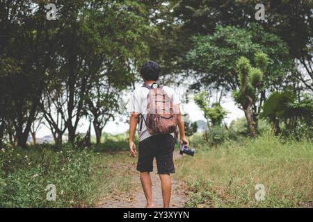 Solo backpacker with camera in hand walks alone. Back view photo of traveler with backpack and camera exploring outdoor, forest, jungle with views of Stock Photo