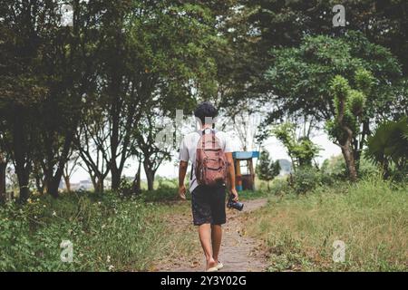 Solo backpacker with camera in hand walks alone. Back view photo of young male with backpack and camera exploring outdoor with trees background Stock Photo