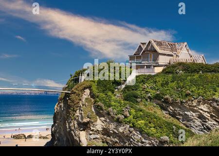 A stunning coastal view featuring a modern house perched on a cliff overlooking a sandy beach. The house has a unique architectural design with a wood Stock Photo