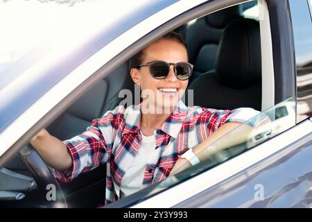 Cheerful young woman in sunglasses driving her new car, enjoying a summer road trip. Stock Photo