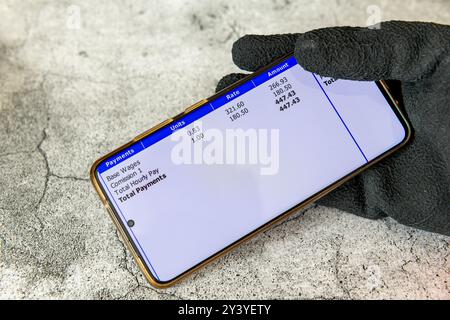 A worker wearing work gloves holding a smart phone with a digital pay slip of wage earnings. Stock Photo