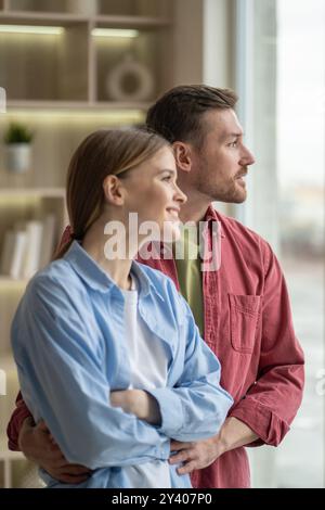 Joyful smiling young family hugging and look out window in snug home. Couple bond, intimacy at home Stock Photo