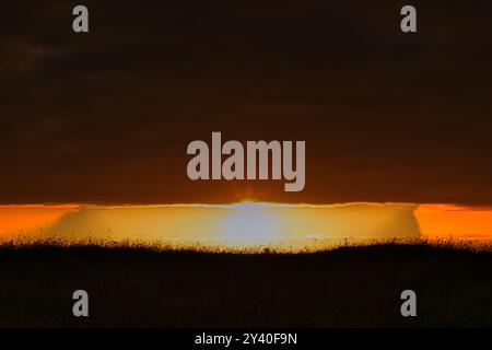 A stunning sunset scene with a dark cloud cover above a glowing horizon. The sun is partially hidden behind the clouds, casting a warm orange and yell Stock Photo