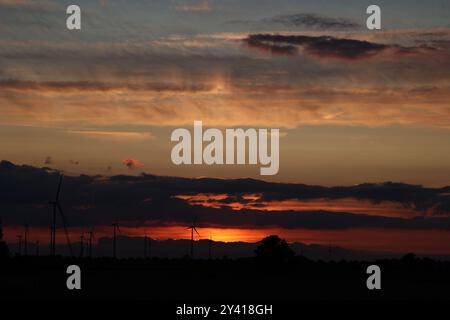 A photo of the glowing sunset with dramatic clouds, wind turbines and trees. Stock Photo