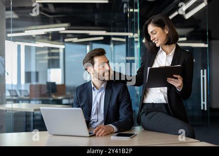 Professional interaction between business colleagues in office expressing teamwork and cooperation. Man with laptop and woman with clipboard showing collaborative work atmosphere Stock Photo