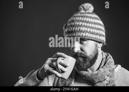 Serious bearded man drinking coffee, cappuccino or tea from cup. Handsome male with beard and mustache in hat and scarf enjoying hot beverage. Fashion Stock Photo