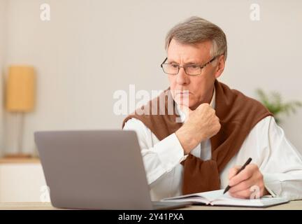 Elderly Man Working On Laptop Taking Notes Sitting In Office Stock Photo