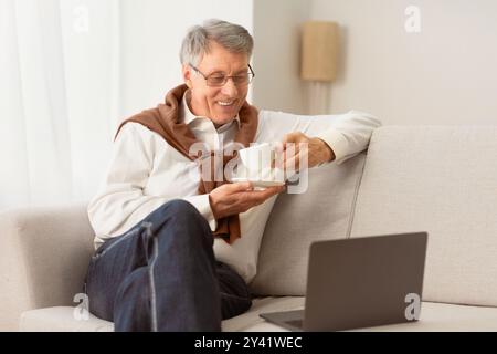 Elderly Man Using Laptop Having Coffee Sitting On Couch Indoor Stock Photo