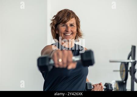elderly woman in sportswear is smiling and punching towards the camera with a dumbbell during a body combat class at the gym. The image highlights her Stock Photo