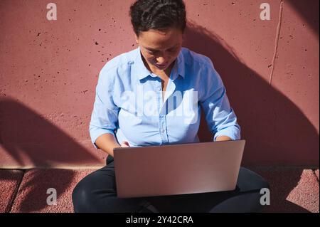 A woman in a blue shirt sits on the ground outdoors, intently using a laptop. She appears to be working or studying, focused on her task in the bright Stock Photo