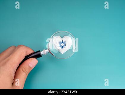 Heart medical check up, health insurance concept. Medical health shield icon on geometric strong white heart, focus in magnifying glass lens, holding Stock Photo