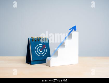 Arrow rise up on minimal white stairway as a growth graph near blue desk calendar with target dart bullseye icon on wooden table and grey blue backgro Stock Photo