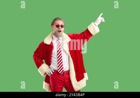 Grey haired surprised man in sunglasses and red Santa style suit pointing on green background. Stock Photo