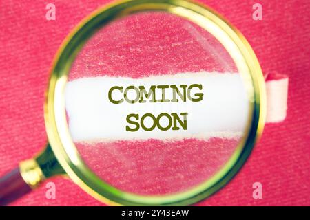 Conceptual coming soon symbol. Copy space. COMING SOON words written on white paper through a magnifying glass under a torn piece of red paper Stock Photo