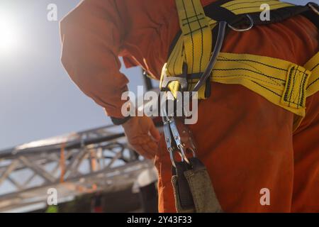 A close-up of a worker wearing a safety harness with a yellow strap, preparing to climb or work at height. The background shows a construction site wi Stock Photo