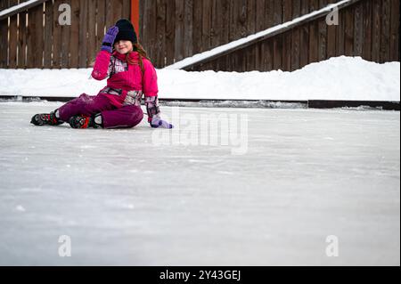 Riga, Latvia - December 29, 2021: A girl in winter clothing sits on the ice rink smiling after falling. Stock Photo