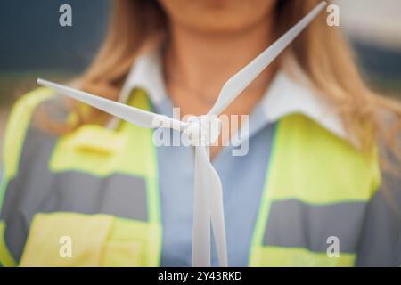 A Female Engineer is proudly Holding a Miniature Wind Turbine Model while standing on a busy Construction Site, highlighting her commitment to Stock Photo