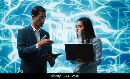 Coworkers solving tasks on laptop and tablet, running artificial intelligence programming scripts. IT staff members using devices to write lines of code for AI applications development, camera A Stock Photo