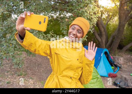 Woman in a yellow jacket is taking a selfie with her cell phone surrounded by nature Stock Photo