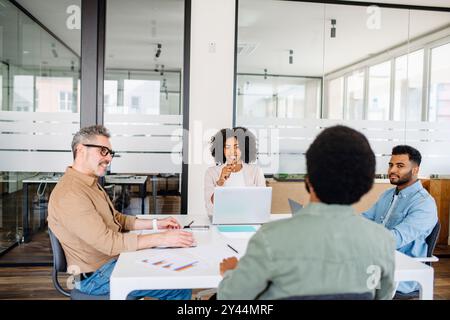 A diverse group of professionals collaborates in a modern office space, with a woman in a white blazer leading the discussion, indicative of a team brainstorming session. Stock Photo