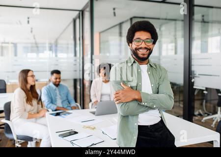 Young man stands with arms crossed in modern office space, showcasing relaxed yet professional work environment, behind him diverse team of colleagues engages in a productive and collaborative meeting Stock Photo