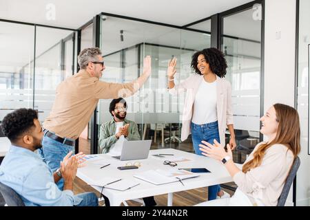 Elation and team spirit are palpable as two colleagues high-five in a contemporary office space, celebrating a collaborative achievement with vibrant enthusiasm. The diverse team applauds happily Stock Photo