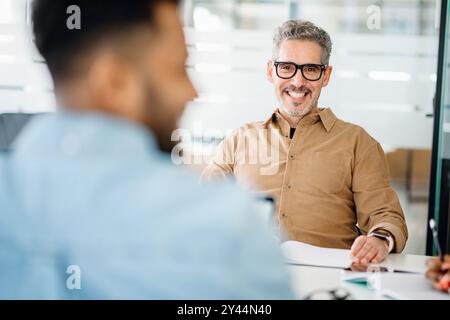 A mature professional man in a tan shirt beams at a colleague out of focus in the foreground, exemplifying a supportive and collaborative office environment during a productive meeting. Stock Photo