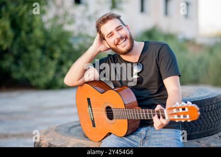 Young man with beard and black t-shirt is playing a classdic guitar and is smilling. He is outdoors and there are some trees and a building behind him Stock Photo