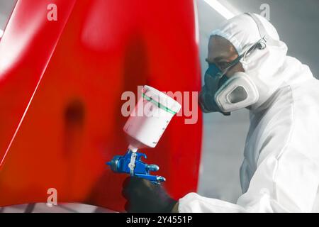 Worker in protective mask and overall spraying base coat of paint on car body elements in painted chamber using professional airbrush. Stock Photo