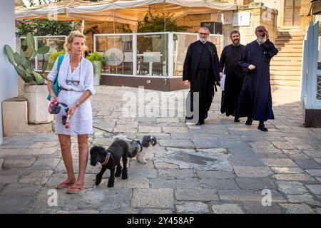 Three Basilian monks in robes walk past a women with two dogs in the ancient city of  Otranto, Apulia, Italy Stock Photo