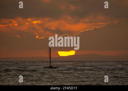 Large, spectacular sun setting at sea, partially hidden by clouds with a floating buoy/ beacon in the foreground. Stock Photo