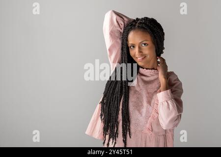 Portrait of smiling young African American female with long braids looking at camera while standing with raised hand behind head against gray backgrou Stock Photo