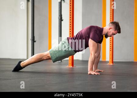 Side view of strong bearded adult male looking down while lying on gym floor and doing calisthenics hand plank hold exercise in daylight Stock Photo