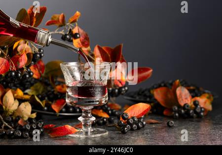 Black chokeberry drink is poured from a bottle into a glass. Sweet drink and fresh berries on a black stone table. Stock Photo