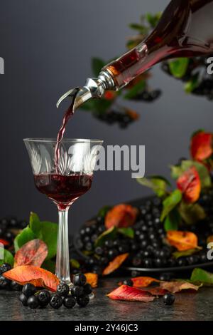 Black chokeberry liqueur is poured from a bottle into a glass. Sweet flavour drink and fresh berries on a black stone table. Stock Photo