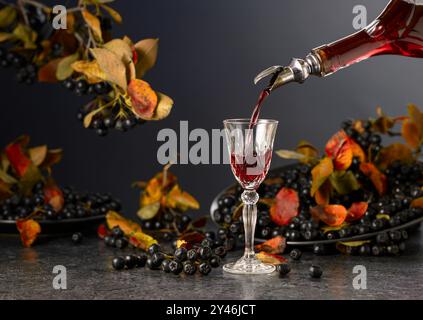 Black chokeberry liqueur and fresh berries on a black stone table. Sweet flavour drink is poured from a bottle into a glass. Stock Photo