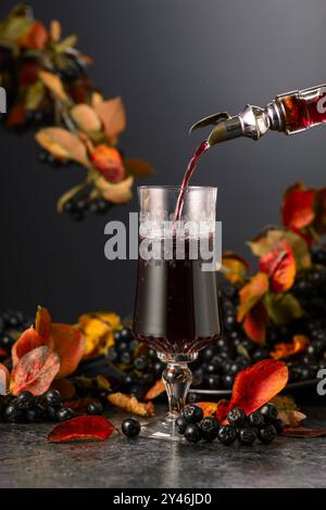 Black chokeberry drink and fresh berries on a black stone table. Sweet  drink is poured from a bottle into a glass. Stock Photo