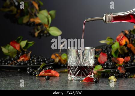 Black chokeberry juice and fresh berries on a black background. Juice or liquor is poured from a bottle into a glass. Stock Photo