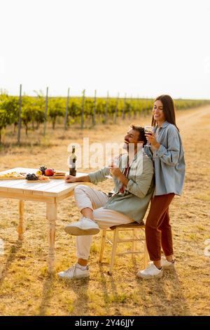 Couple relaxes at a rustic table in a sunlit vineyard, savoring wine and gourmet snacks. They share laughter and joy, surrounded by lush vines and the Stock Photo