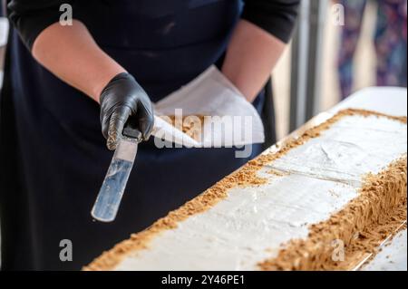 A baker wearing black gloves spreads crumbs around the edges of a white cake. Shallow depth of feld Stock Photo