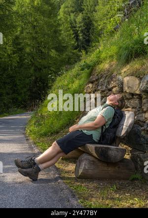 Tired middle aged woman with backpack leaned back on bench and closed her eyes on trail with alpine mountain peaks.   Grossglockner. Hohe Tauern Natio Stock Photo