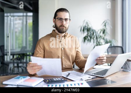 Business professional focuses on paperwork at office desk, featuring laptop, charts, and calculator. Modern workspace suggests analysis, planning, and productivity in financial or corporate setting. Stock Photo