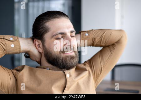Man with beard enjoying moment of relaxation in office setting. Hands behind head, expressing comfort and satisfaction. Casual shirt, confident expression symbolizes stress-free atmosphere Stock Photo