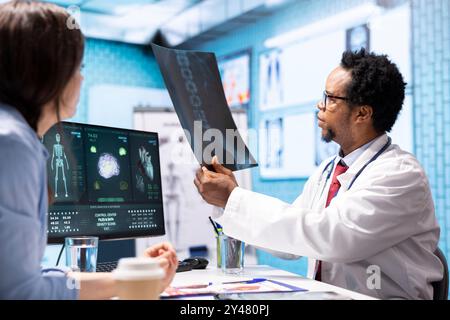 Professional medic analyzing x ray imaging and test findings in office, giving the diagnostic report for female patient at check up. African american specialist examines scans for interpretation. Stock Photo