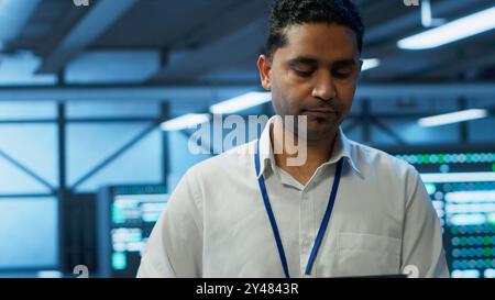 Engineer walking in data center, monitoring security threats, implementing cybersecurity measures. Indian man ensuring compliance with regulations to protect data center gear, camera A Stock Photo