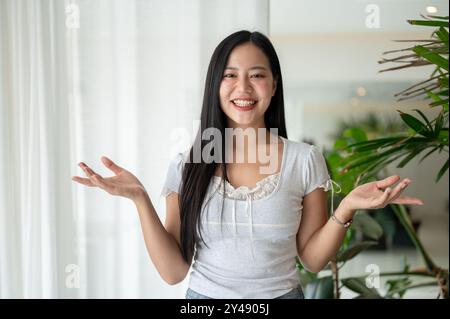 A beautiful, cheerful Asian woman is standing by a window in a room, smiling at the camera and holding her palm open, posing as if recommending someth Stock Photo